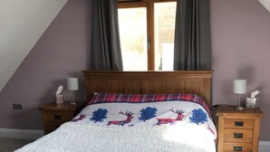 A bedroom with a sloped ceiling, a wooden bed frame, a plaid bedspread, two bedside tables with lamps, and a window with curtains. - Tigh a Chabar, Savary. Nr Lochaline.  Cosy cottage, sleeps 4.  Sea views. (Morvern, Argyll)