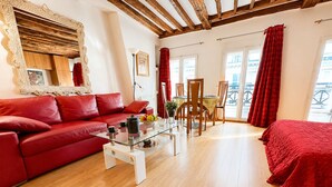 Ein Zimmer mit einem roten Sofa, einem Glastisch, einem Holzespeisetisch und Stühlen. Es gibt einen Balkon mit roten Vorhängen und Blick ins Freie.