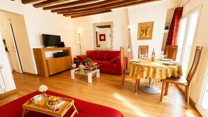 Un salon chaleureux avec un canapé rouge, un téléviseur, un meuble en bois et un coin repas avec une table mise pour deux personnes.