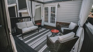 Ein überdachter Patio-Bereich mit Korbmöbeln, einem Streifen Teppich und einer Glastür.