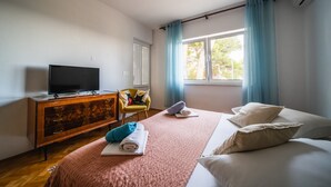Hotellihuone, jossa on sänky, puinen tv-taso, sinisillä verhoilla varustettu ikkuna ja näkymä puita kohti.