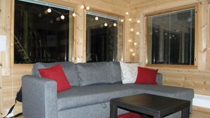 Una habitación acogedora con un sofá gris, una mesita pequeña y una alfombra roja. Hay ventanas amplias con luces de adorno y una persiana.
