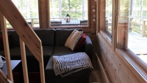 Interior de una cabaña de madera con un sofá, una ventana que da a un bosque y una escalera.