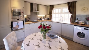 A modern kitchen with a microwave, oven, and washing machine. There's a table with a vase of flowers and a clock on the wall. - West Mead Cottage (Wirral)