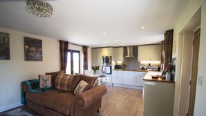 A living room with a brown sofa, a dining area, and a kitchen with white cabinets. - West Mead Cottage (Wirral)