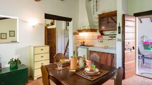 Un coin repas rustique avec une table en bois dressée pour deux personnes, un mur en briques et un espace cuisine visible au fond.