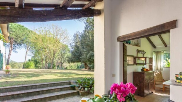 Un patio couvert avec vue sur un jardin, une table sur laquelle se trouve un plateau avec des tasses vertes et une plante en pot, et un aperçu d’une cuisine.