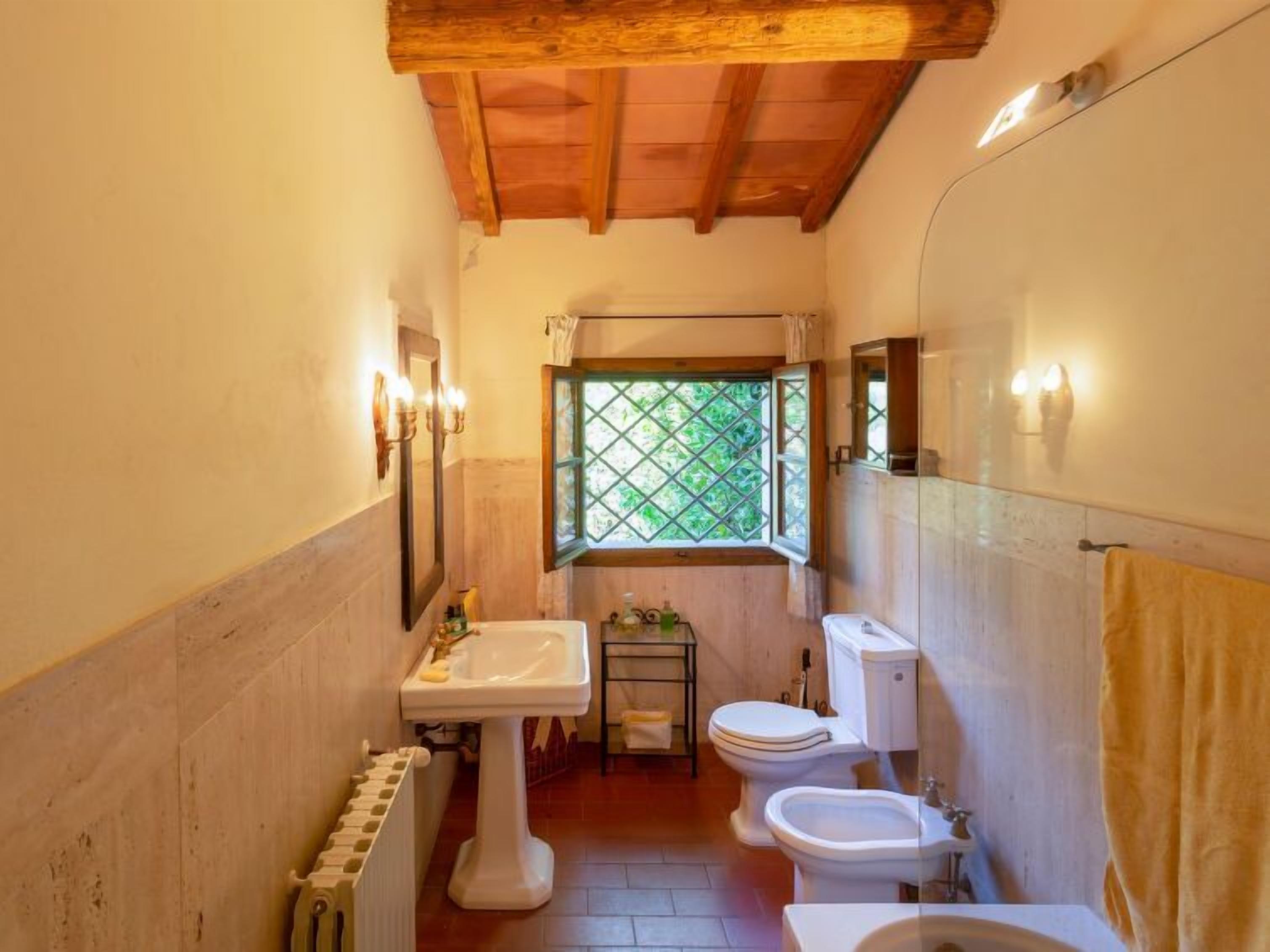 A compact bathroom with a wooden ceiling, a window with a green lattice, a pedestal sink, two toilets, and a bidet.