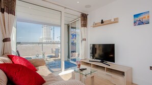 Ett modernt vardagsrum med en stor platt-TV, ett soffbord i glas och en soffa med röda kuddar. Det finns en skjutdörr i glas som leder ut till en balkong.