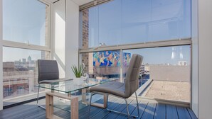 En modern balkong med ett glasbord och två stolar, en krukväxt och ett stort fönster med utsikt över stadsbilden.