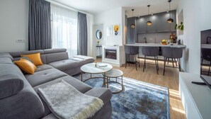 Een moderne woonkamer met een grijze bank, een salontafel en een blauw vloerkleed. Er is een keuken met grijze kastjes en hanglampen.