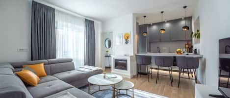 A modern living room with a grey sofa, a coffee table, and a blue rug. There is a kitchen area with grey cabinets and pendant lights.