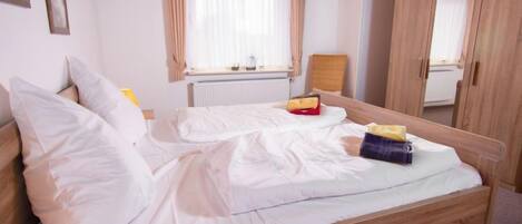A bedroom with a wooden bed, white bedding, a bedside table with a lamp, a window with curtains, and a wardrobe.