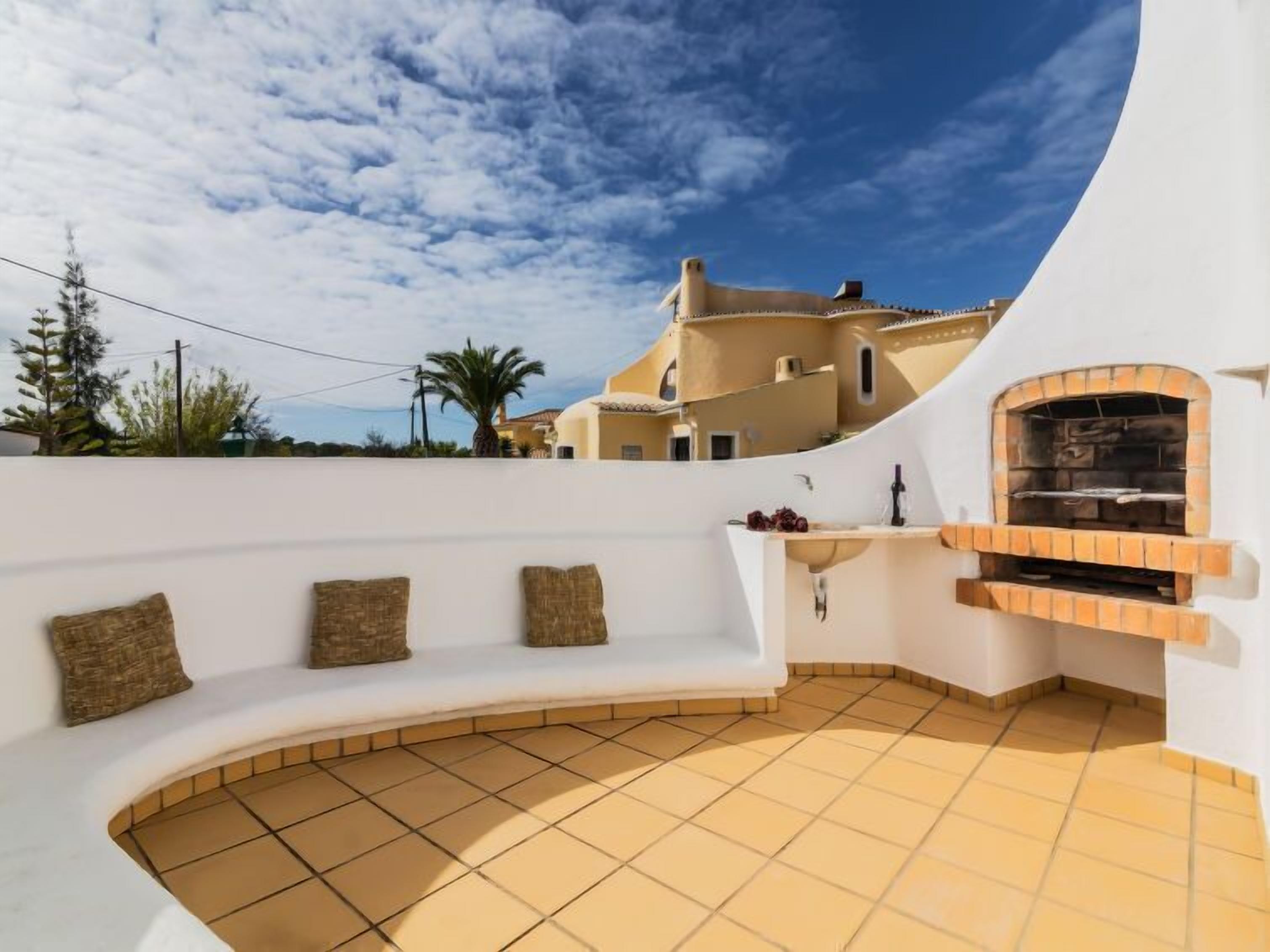 A rooftop terrace with a built-in oven, a white wall with three cushions, and a view of a yellow building under a blue sky.