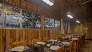 A dining area with wooden walls and large windows, featuring a long table set for multiple guests. - Hotel Nirala (Darjeeling)