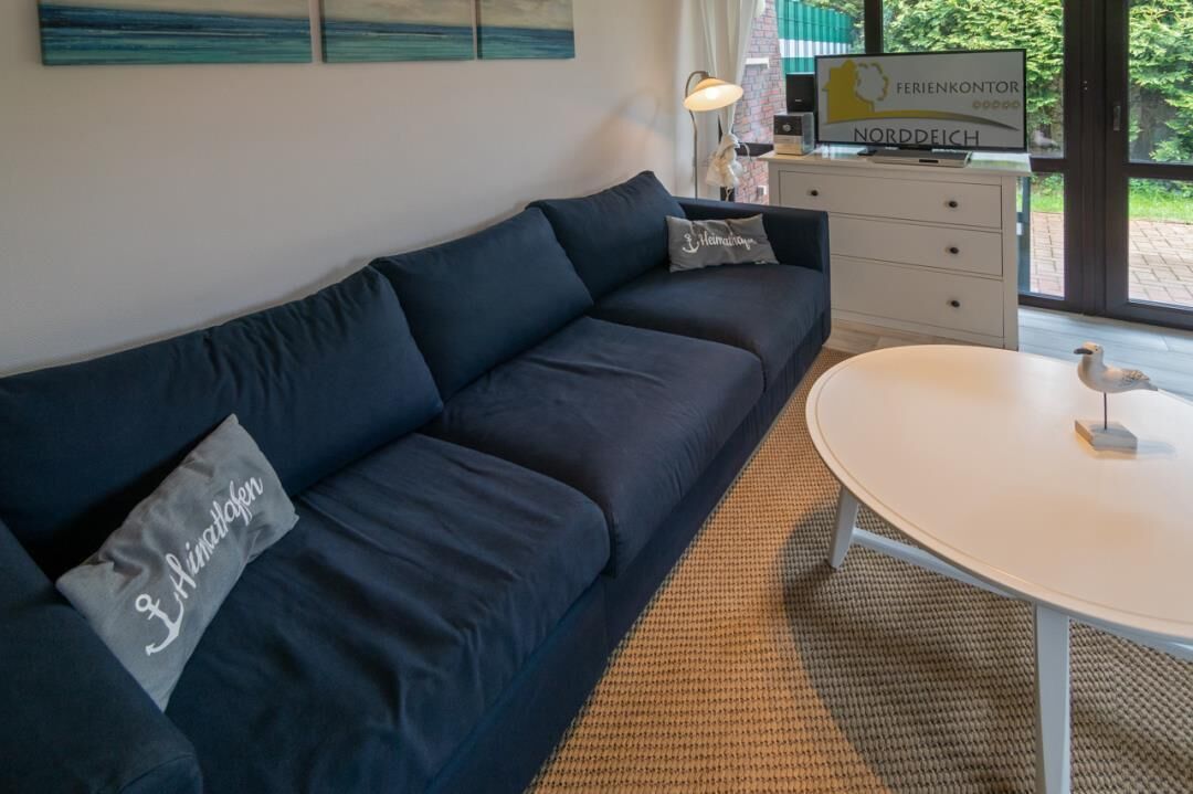 Ein modernes Wohnzimmer mit einem dunkelblauen Sofa, einem runden weißen Tisch und einem dekorativen Vogel auf dem Tisch. Ein Fernsehgerät steht auf einer weißen Kommode, und ein Fenster bietet Blick ins Grüne.