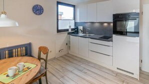 Une cuisine compacte avec des meubles blancs, un four encastré, une fenêtre et un coin repas comprenant une table et des chaises en bois.