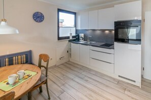 Une cuisine compacte avec des meubles blancs, un four encastré, une fenêtre et un coin repas comprenant une table et des chaises en bois.