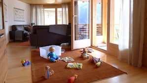 A cozy living room with a wooden ceiling, a brown sofa, a child playing with toys, and a large window offering a view of the outdoors. - Apartment Gloria by Interhome (Mürren)