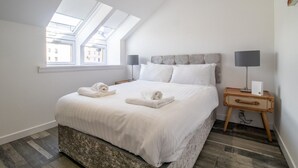 Une chambre avec une lucarne, un lit recouvert de draps blancs, une table de chevet avec une lampe et une valise.