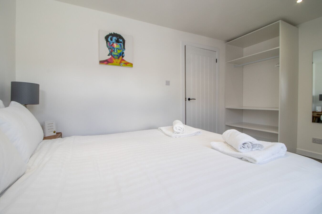 A hotel room with a bed, a painting on the wall, a wardrobe, and a nightstand with a lamp.