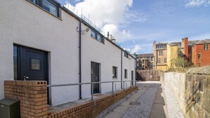 Un bâtiment à l’extérieur blanc, avec une porte noire et une petite fenêtre. Un mur de briques se trouve devant le bâtiment, et un chemin de gravier y mène.
