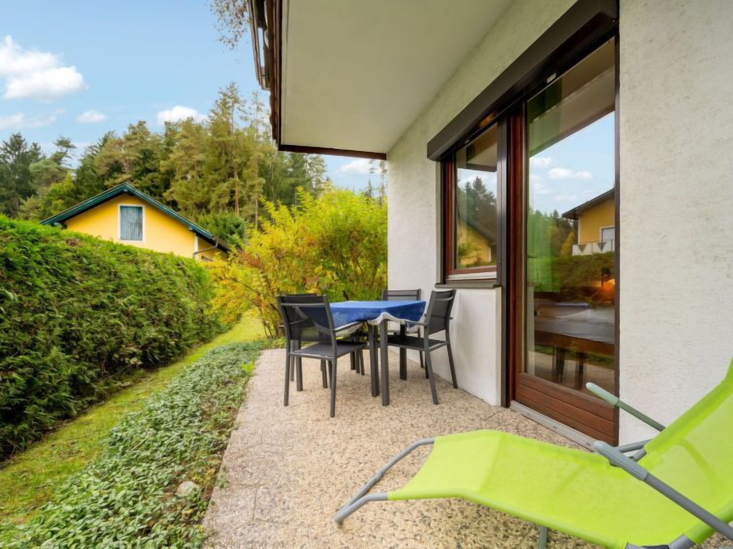 Eine Terrasse mit Tisch und Stühlen, ein grüner Sessel und Blick auf ein Haus und Bäume.