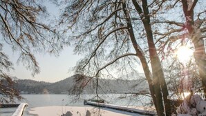 Schneebedeckte Landschaft mit Holzlattenzaun, Bäumen und einem Gewässer.