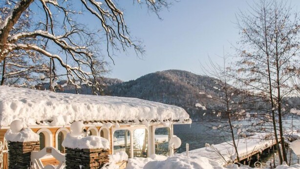 Ein schneebedecktes Chalet mit Steinkamin, Holzbalken und Blick auf einen See und Berge.