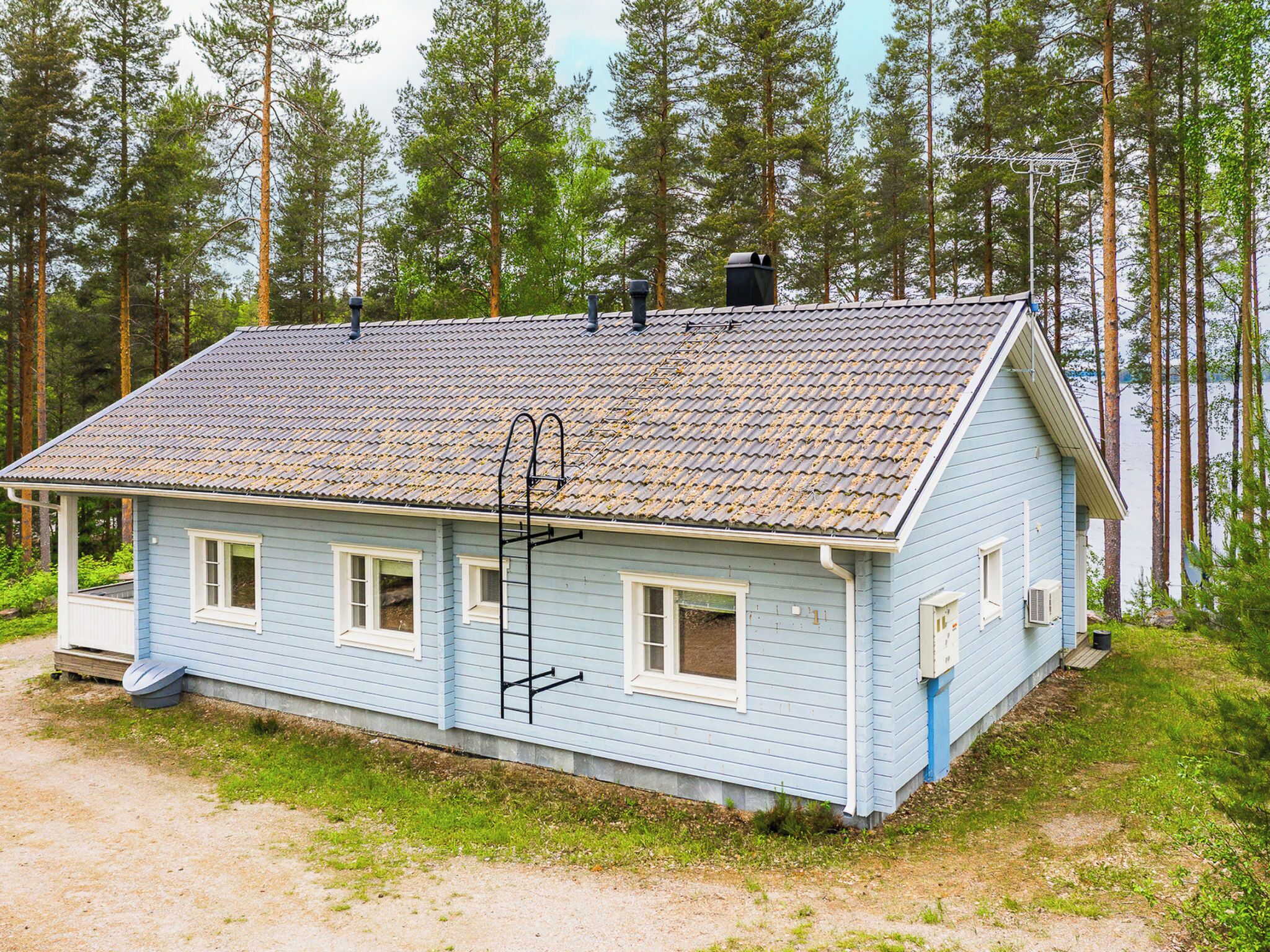 Une maison bleu clair avec un toit en tuiles, entourée de grands arbres et bordée par un plan d’eau.
