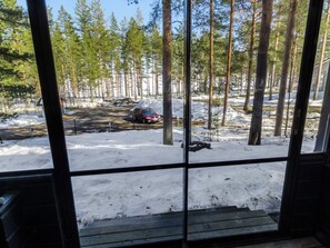 Une vue à travers une fenêtre donnant sur une terrasse en bois, un sol recouvert de neige et une forêt d’arbres imposants.