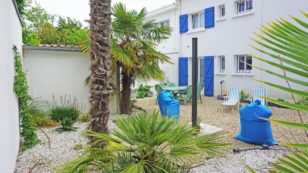 A courtyard with a palm tree, a small garden, and a white building with blue shutters. There are two chairs and a table in the center.