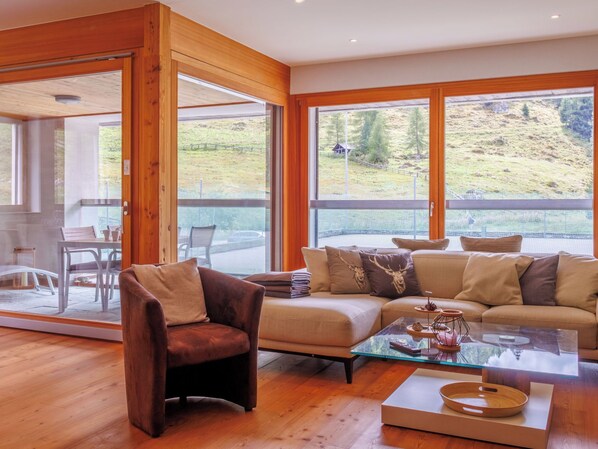 Una sala de estar moderna con una mesa de centro de vidrio, un sofá y una silla. Hay un ventanal que ofrece vistas a un paisaje cubierto de hierba.