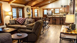 A cozy living room with a sofa, armchairs, and a coffee table. There is a dining area with a table and chairs, and a kitchen with a refrigerator and cabinets.