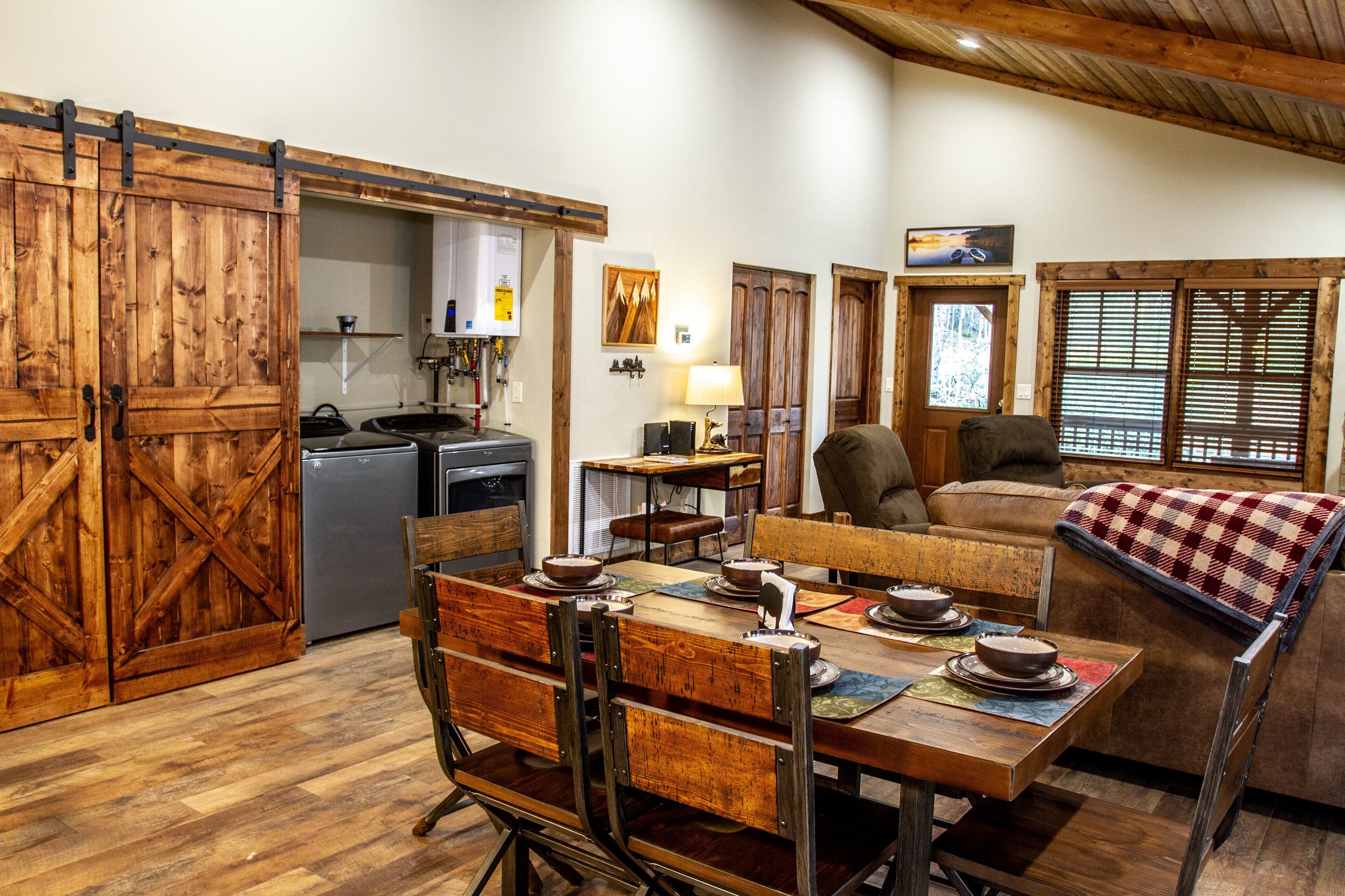 A rustic interior with a wooden dining table set for four, a sofa, and a kitchen area with a refrigerator and stove.