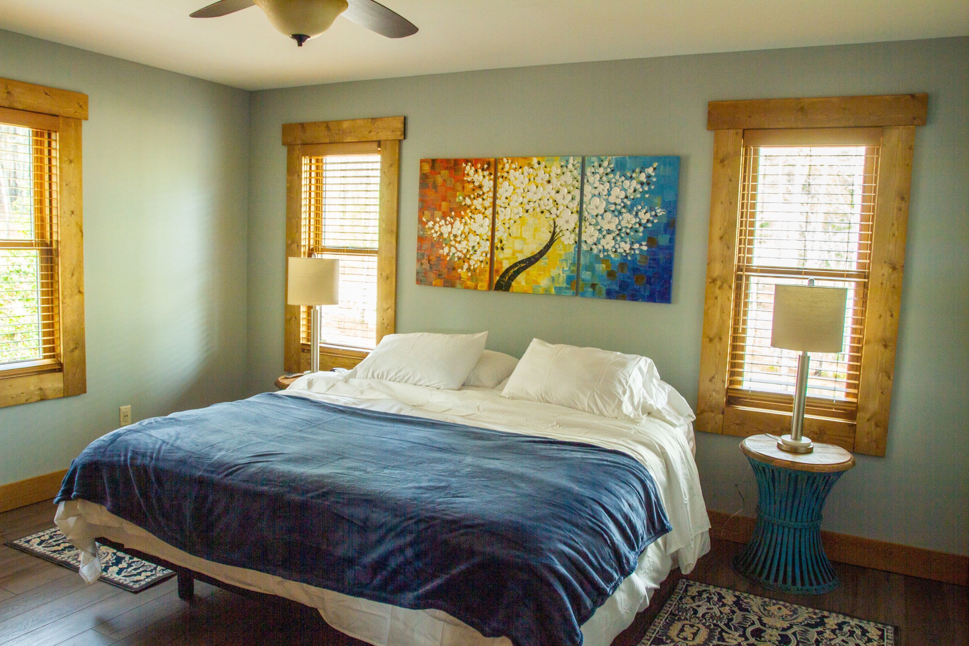A bedroom with a bed, a painting, two windows with blinds, and a ceiling fan.