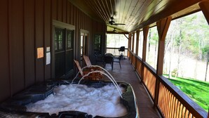 A covered outdoor hot tub area with a wooden deck, chairs, and a view of a green landscape.
