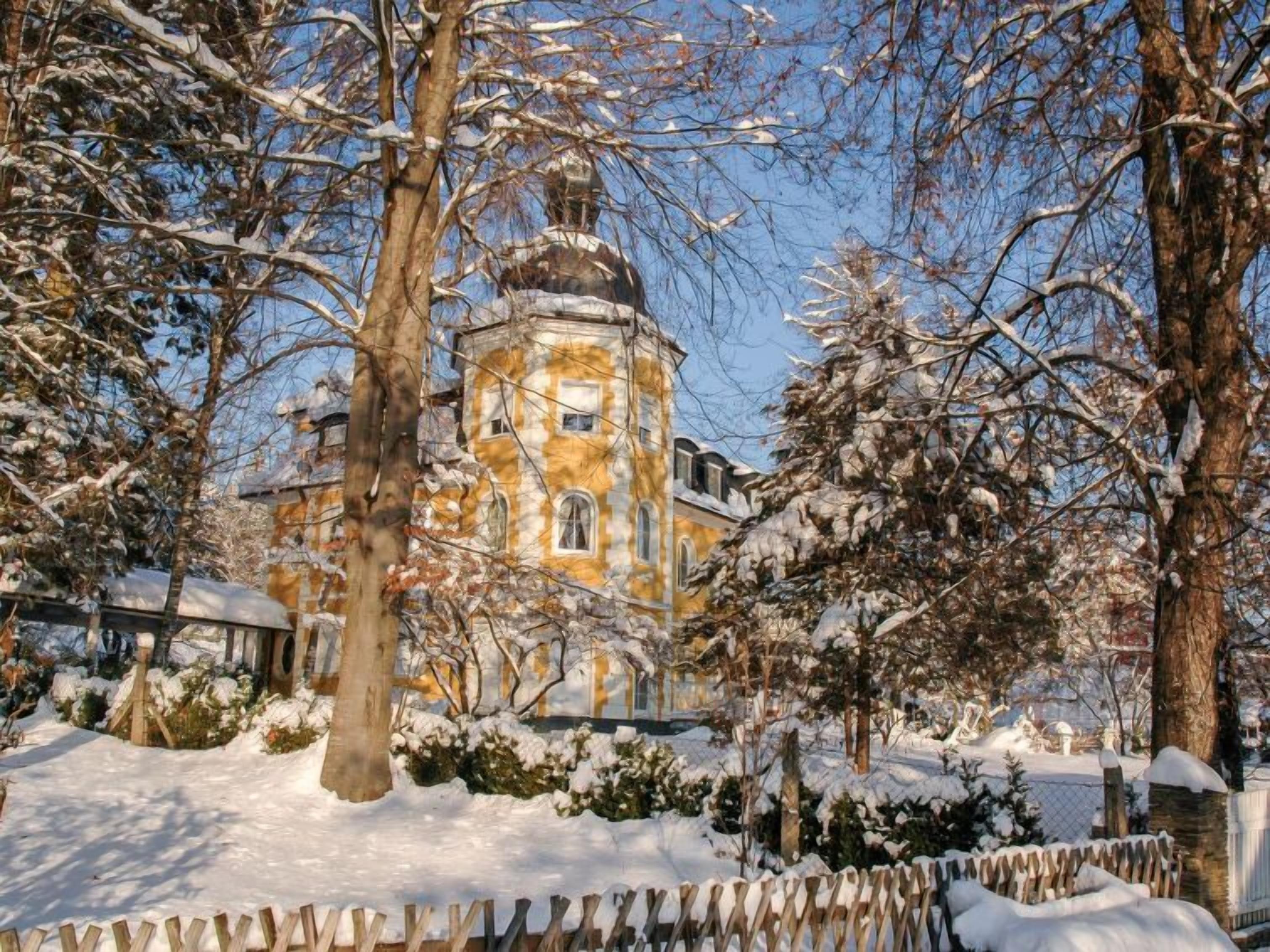 A historic building with a central tower, surrounded by snow-covered trees and a wooden fence.