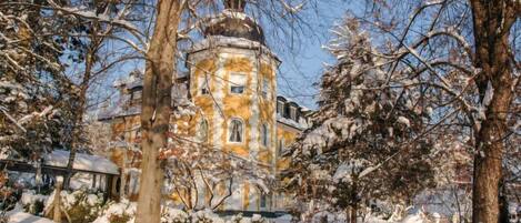 A historic building with a central tower, surrounded by snow-covered trees and a wooden fence.