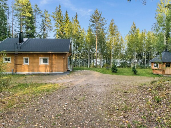 A wooden cabin with a gravel driveway, surrounded by a forest with tall trees.