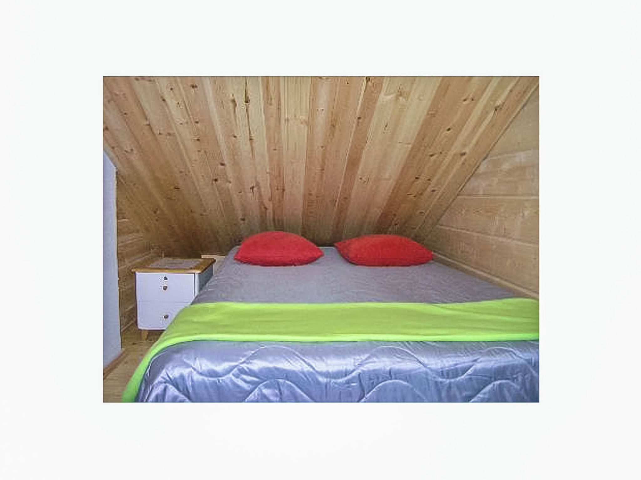 Une petite pièce aux murs lambrissés, avec un lit, deux oreillers rouges, une couvre-lit verte et argentée, et une table de chevet blanche sur laquelle se trouve une boîte.