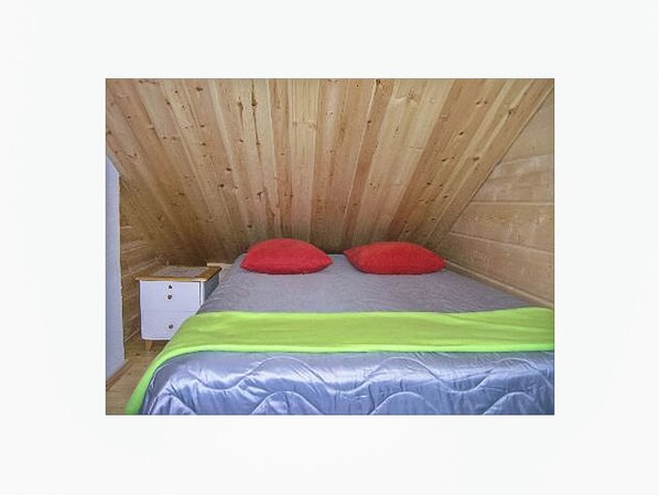 A small, wooden-paneled room with a bed, two red pillows, a green and silver bedspread, and a white nightstand with a box on it.