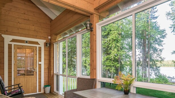 A wooden cabin with a covered porch, wicker furniture, and large windows offering a view of the forest and lake. - Villa eerola by Interhome (Juva)
