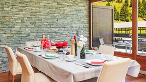 Una mesa de comedor dispuesta con platos, cubiertos y vasos, rodeada de sillas, frente a una pared de piedra con vistas a la vegetación.