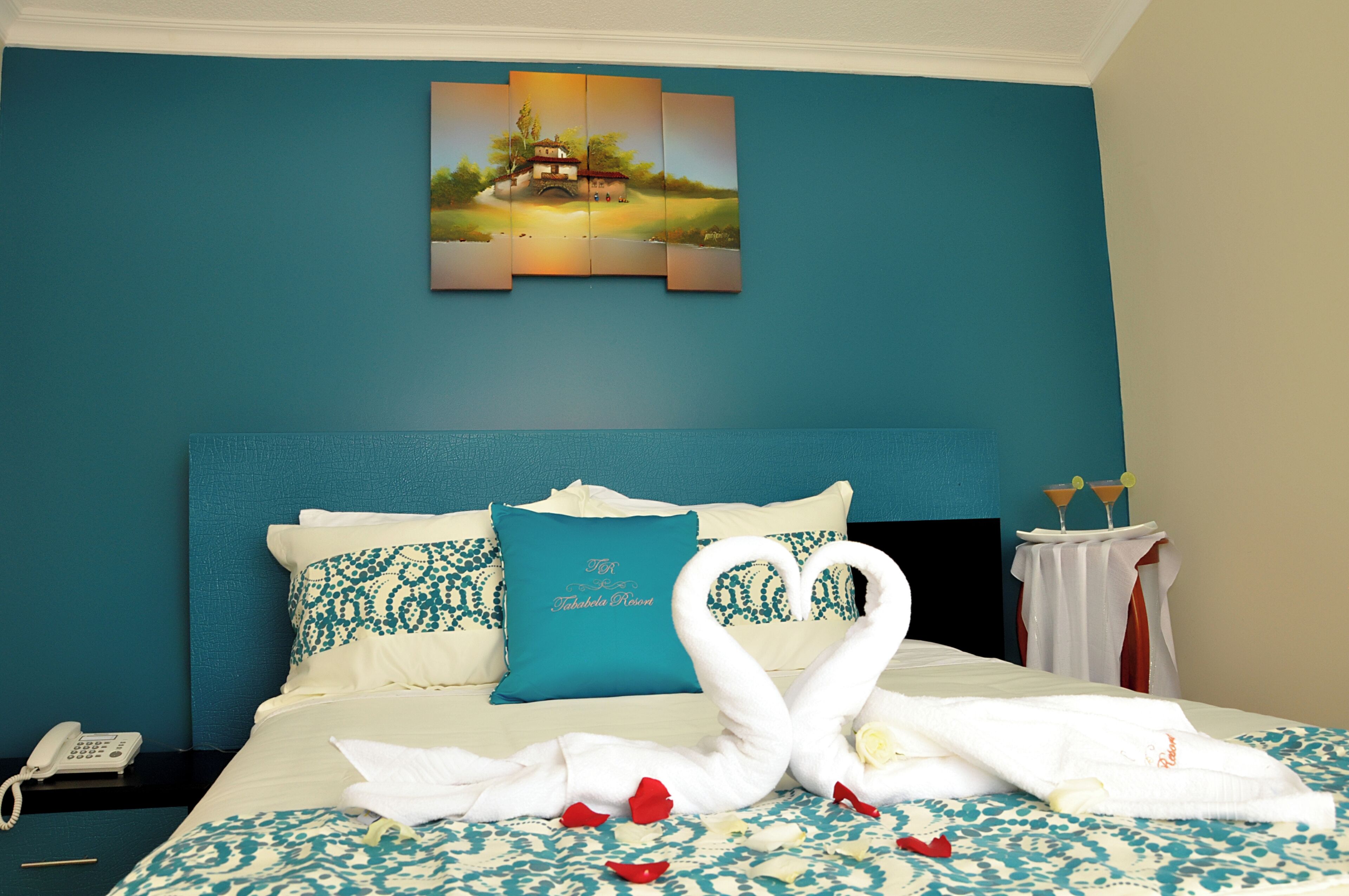 A hotel room with a teal wall, a bed with a blue and white patterned bedspread, a teal pillow, and a painting above the bed.