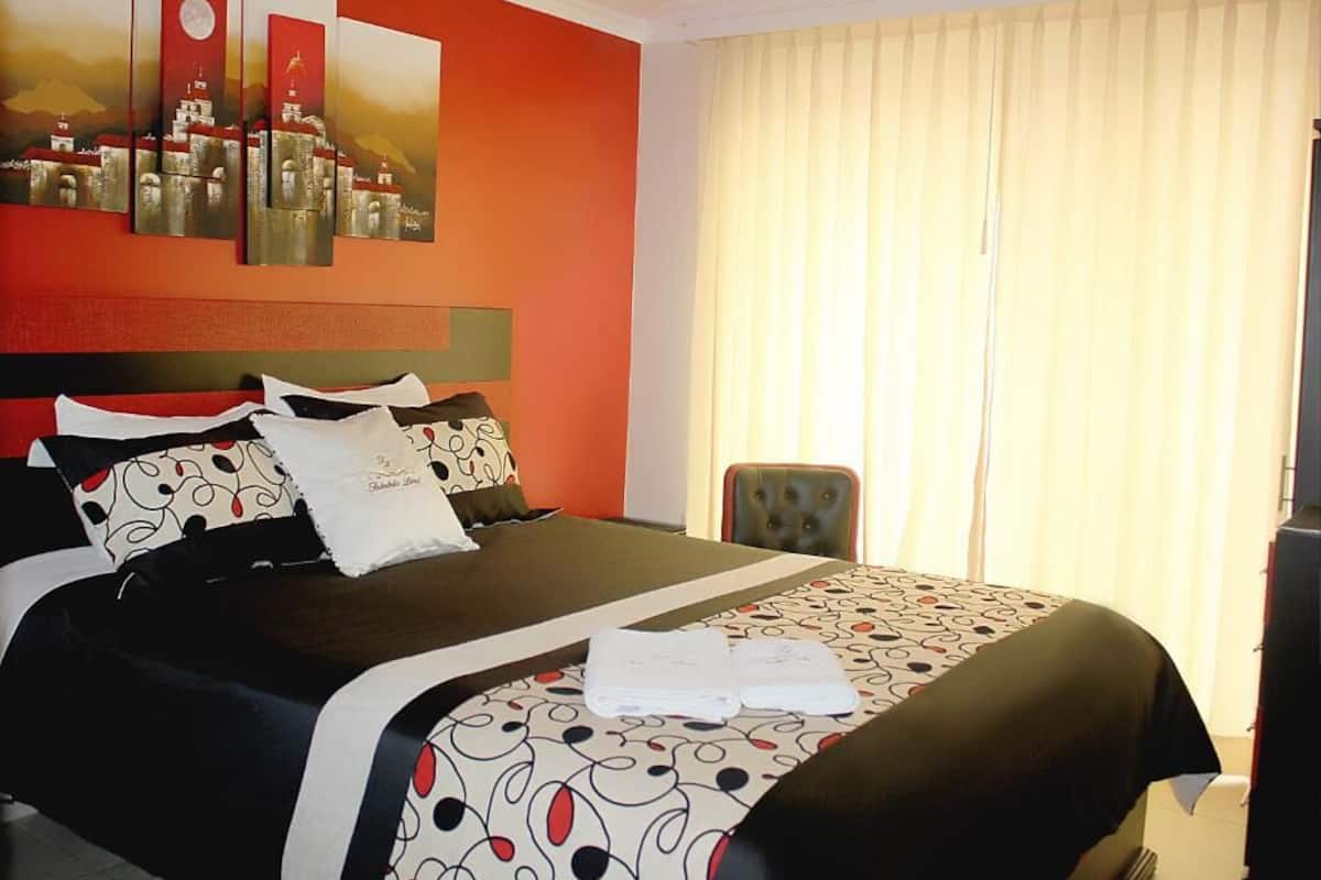 A hotel room with a bed, a headboard, a window with curtains, and a painting on the wall.