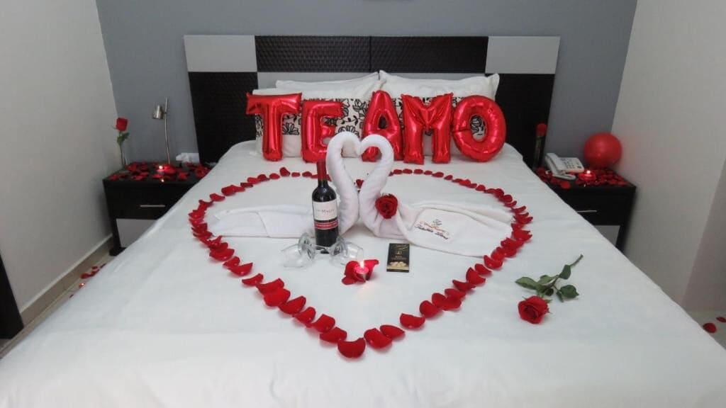 A bed with a heart-shaped arrangement of red rose petals, a bottle of wine, and a swan-shaped decoration.