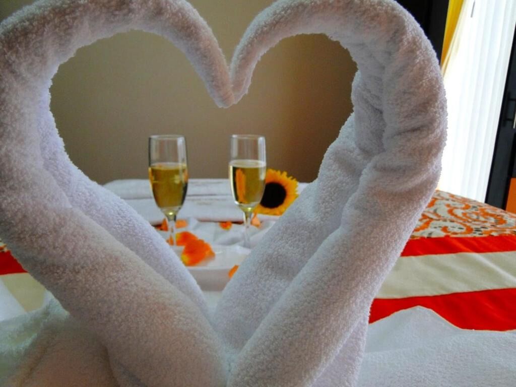 A bed with a heart-shaped towel arrangement, two glasses of champagne, and a sunflower on the bed.