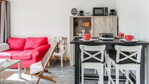 A compact living space with a red sofa, a dining table with chairs, a kitchen area with a microwave, and a small seating area with a chair and a table. - Pegase Phenix by Interhome (Le Corbier)