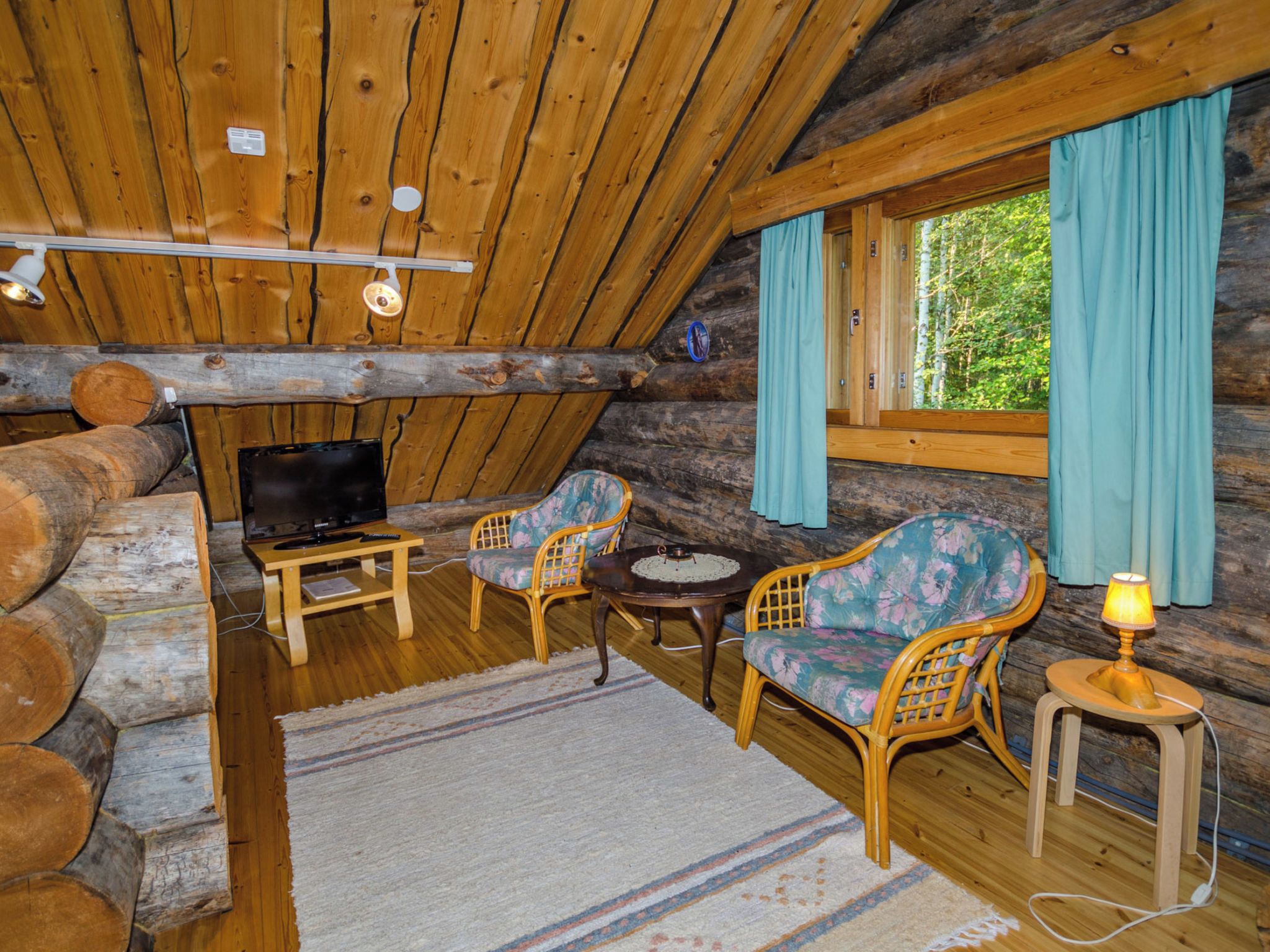 Interior de cabana de madeira com televisão, duas cadeiras de vime, uma mesa pequena e uma janela com cortinas.